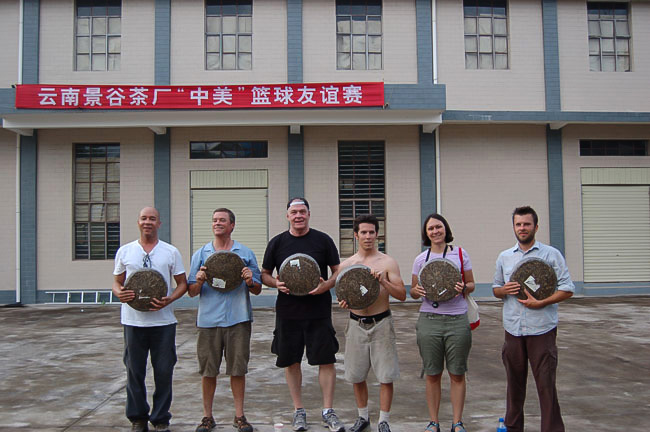 Six people holding large puer cakes on the court in front of a red banner.