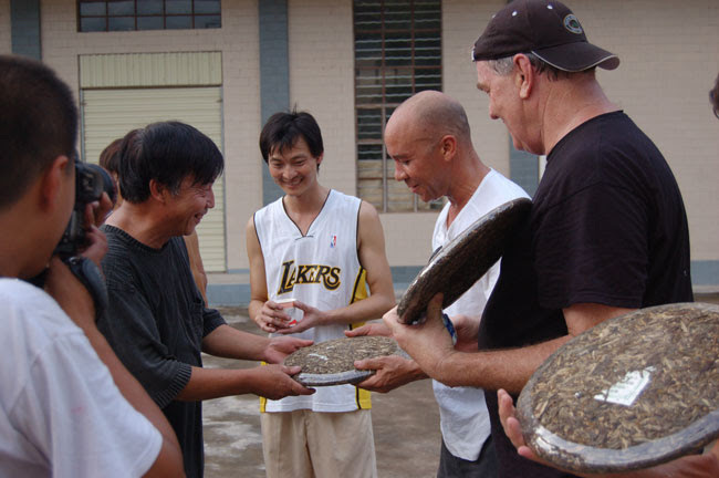 A smiling man passing a large round puer cake to Austin, surrounded by other factory team members and tea tour members.