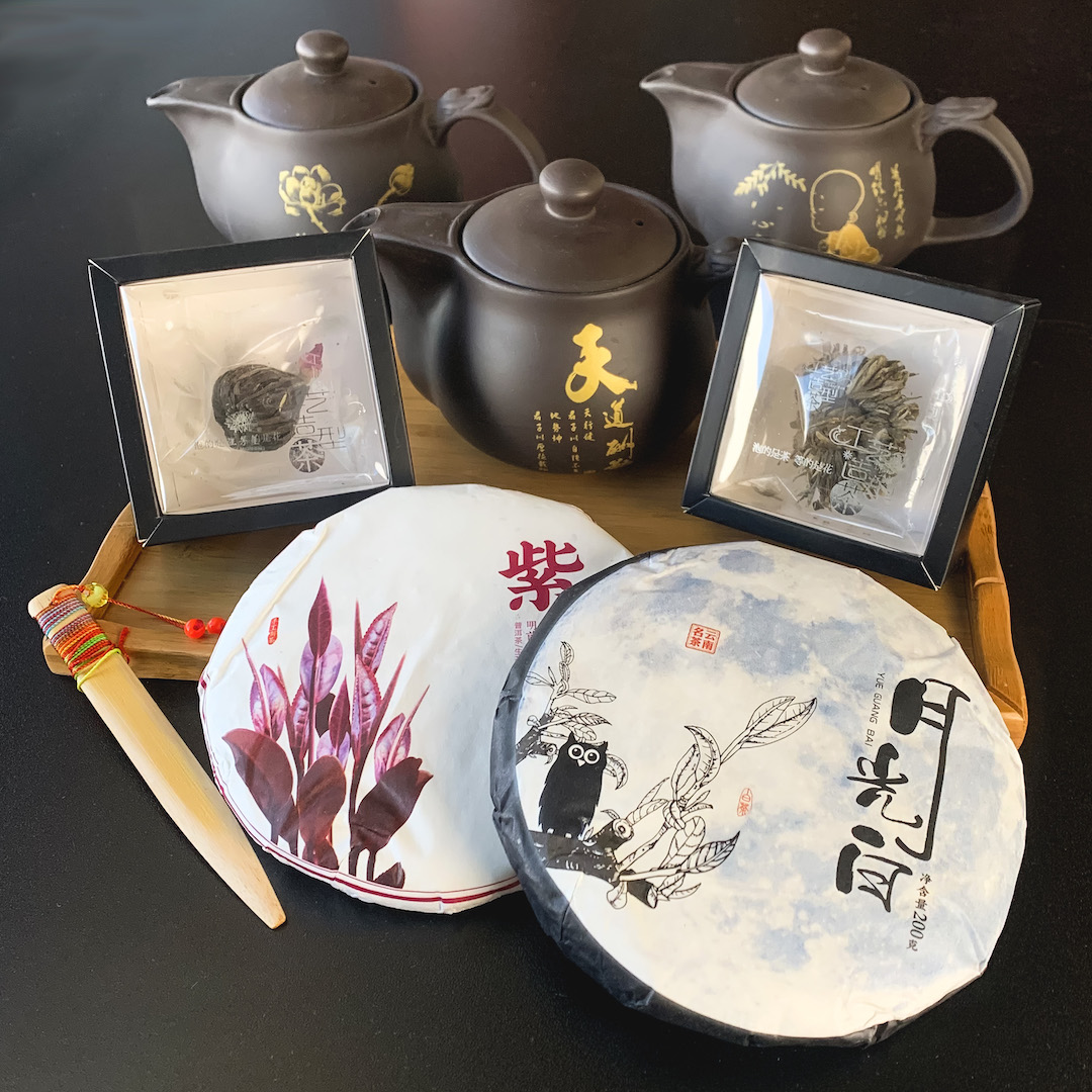 Two small paper-wrapped puer cakes, two blooming teas in individual boxes, and three yixing teapots gathered together on and around a bamboo tray.