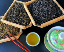 Three bamboo trays of dry black tea arranged in a fan next to an ornate enameled green gaiwan, a set of wooden tea tweezers, and a small porcelain cup of brewed tea.