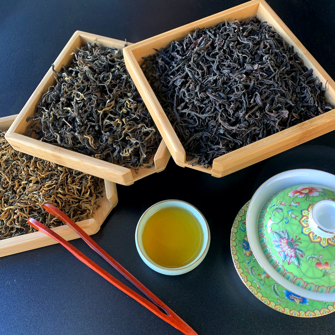 Three bamboo trays of dry black tea arranged in a fan next to an ornate enameled green gaiwan, a set of wooden tea tweezers, and a small porcelain cup of brewed tea.