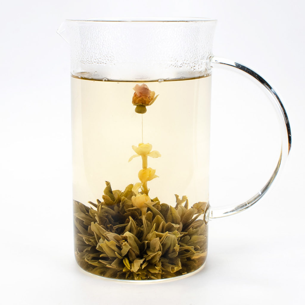 A full glass pitcher holding a fully bloomed ball of green tea with a chain of jasmine flowers topped with a single rose bud springing from the center.