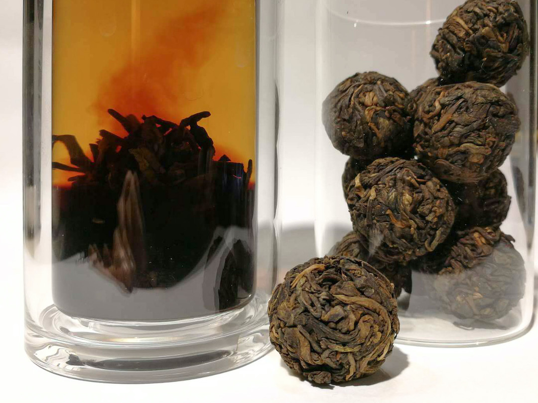 A small shu puer ball set in front of a jar of others and a tall glass filled with water and another ball of tea unfurling in the bottom, with infused tea rising from it in a darker plume.