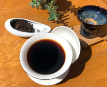 In bright sunlight, a large white gaiwan full of very dark red-brown shu puer tea, set on a worn wooden surface next to a ceramic tea strainer and display dish of dry tea leaves. The leaves of a small green succulent plant poke in from the top of the frame.