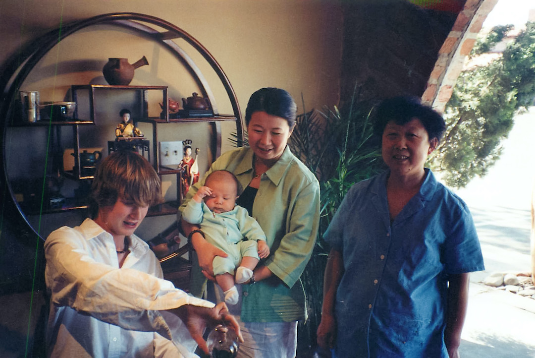Two women standing in the teahouse, one holding a baby, while a young man seated next to them pours tea.