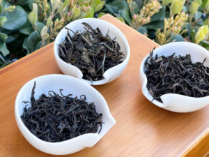Three porcelain dishes full of dark dry tea leaves, one a long thin and twisted black tea, one fluffy and dark green with larger leaves and some buds, and one a broader dark twisted rock wulong.