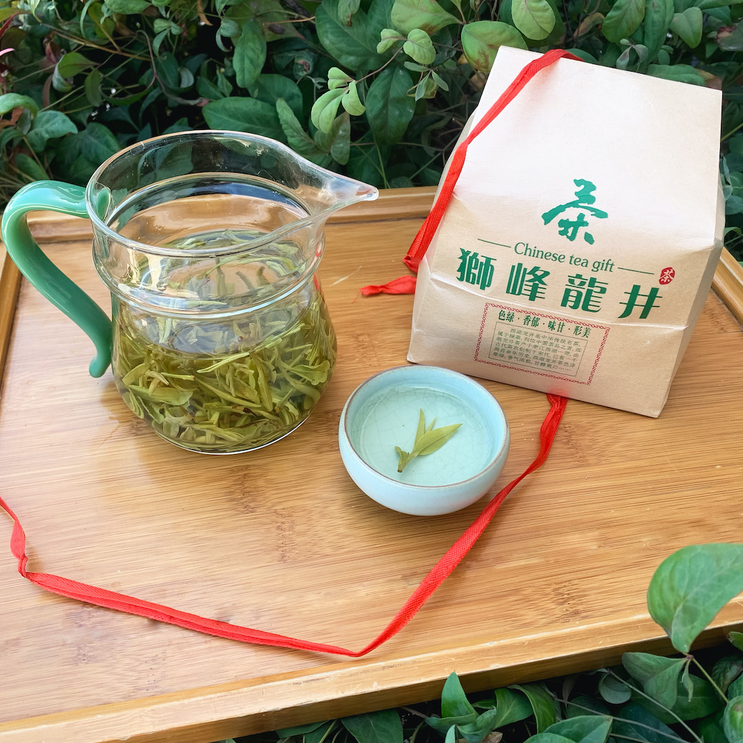 A glass pitcher of brewing green tea, a small filled teacup with a single tea sprig floating in the middle, and a small brown paper package of tea leaves with a trailing untied red ribbon around it, all on a bamboo tray surrounded by green and red bamboo-like leaves of a plant.