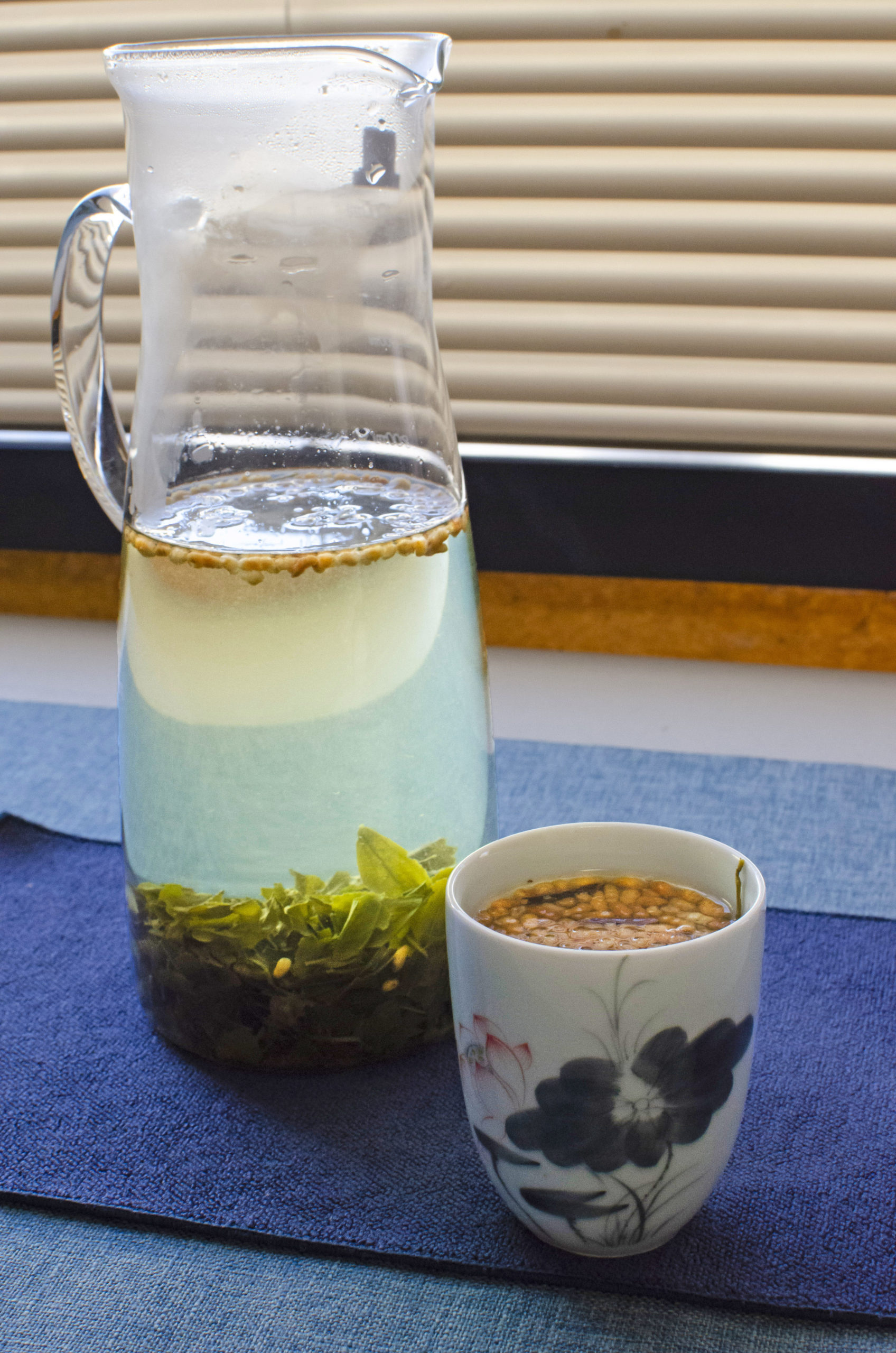 A glass pitcher full of green tea leaves and toasted rice sits on a blue surface infront of a window.
