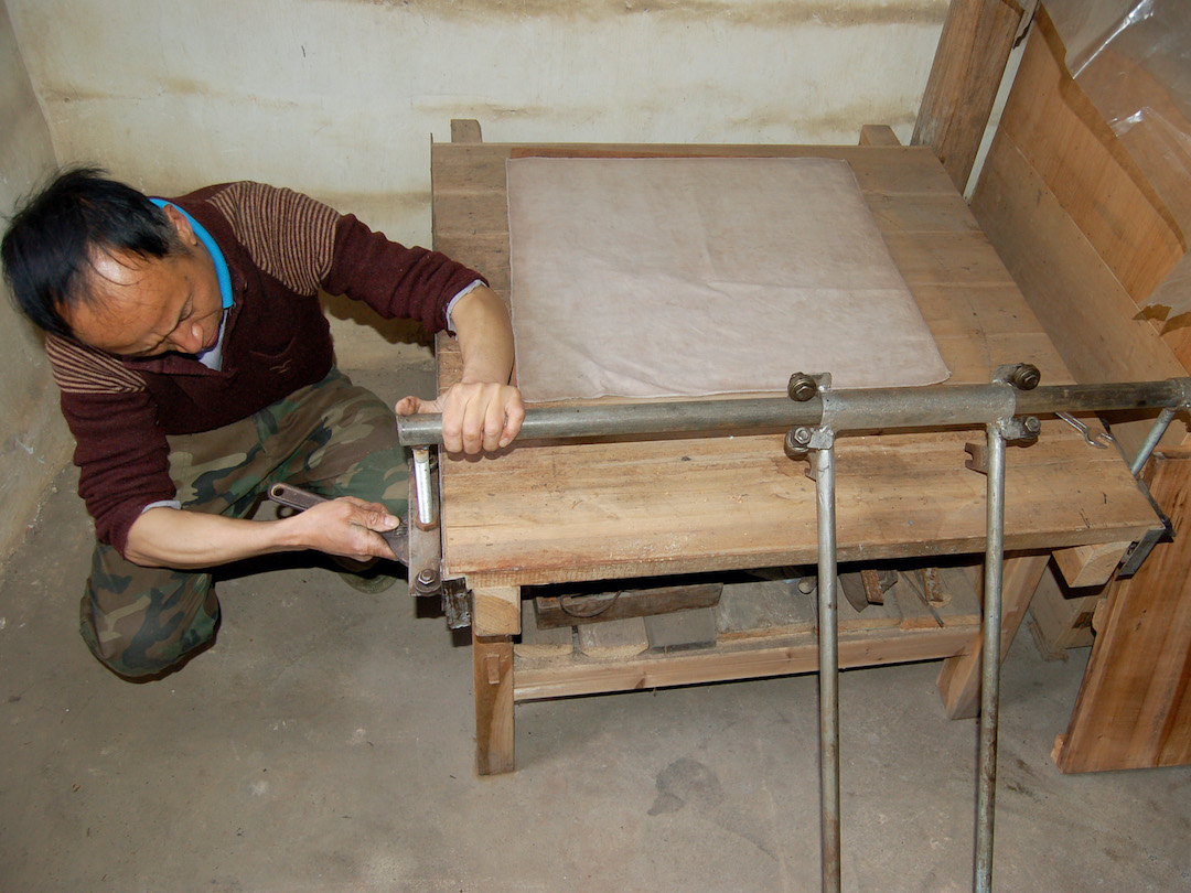 A man kneeling next to the manual press machine for flattening Tai Ping Houkui green tea, a wooden bench with metal bar attachments. He is tightening a bolt.