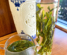 Brewing two green teas in a tall glass bottle filled with very long green leaves and a round squat pitcher with rounded small single leaves dancing in it, set on a wooden tray next to a classical white and blue Chinese vase.