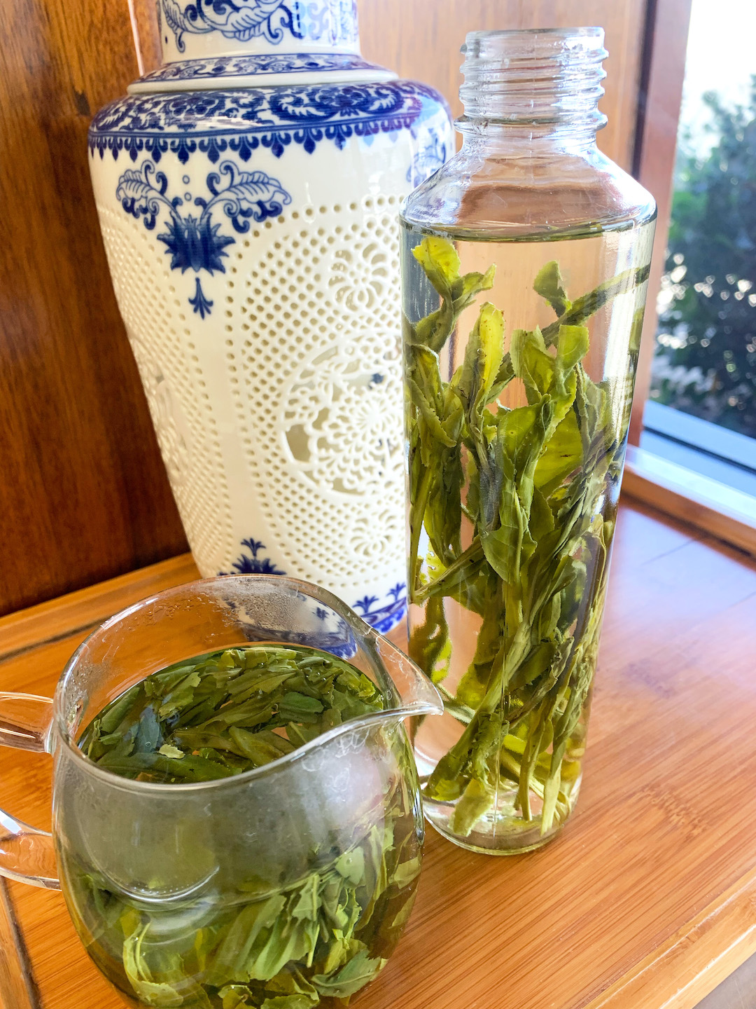Brewing two green teas in a tall glass bottle filled with very long green leaves and a round squat pitcher with rounded small single leaves dancing in it, set on a wooden tray next to a classical white and blue Chinese vase.