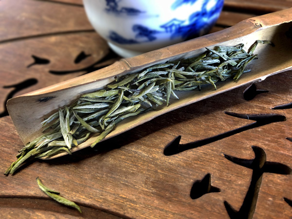 Slender green and silver tea buds in a bamboo scoop sit next to a blue and white tea cup on a wooden table.