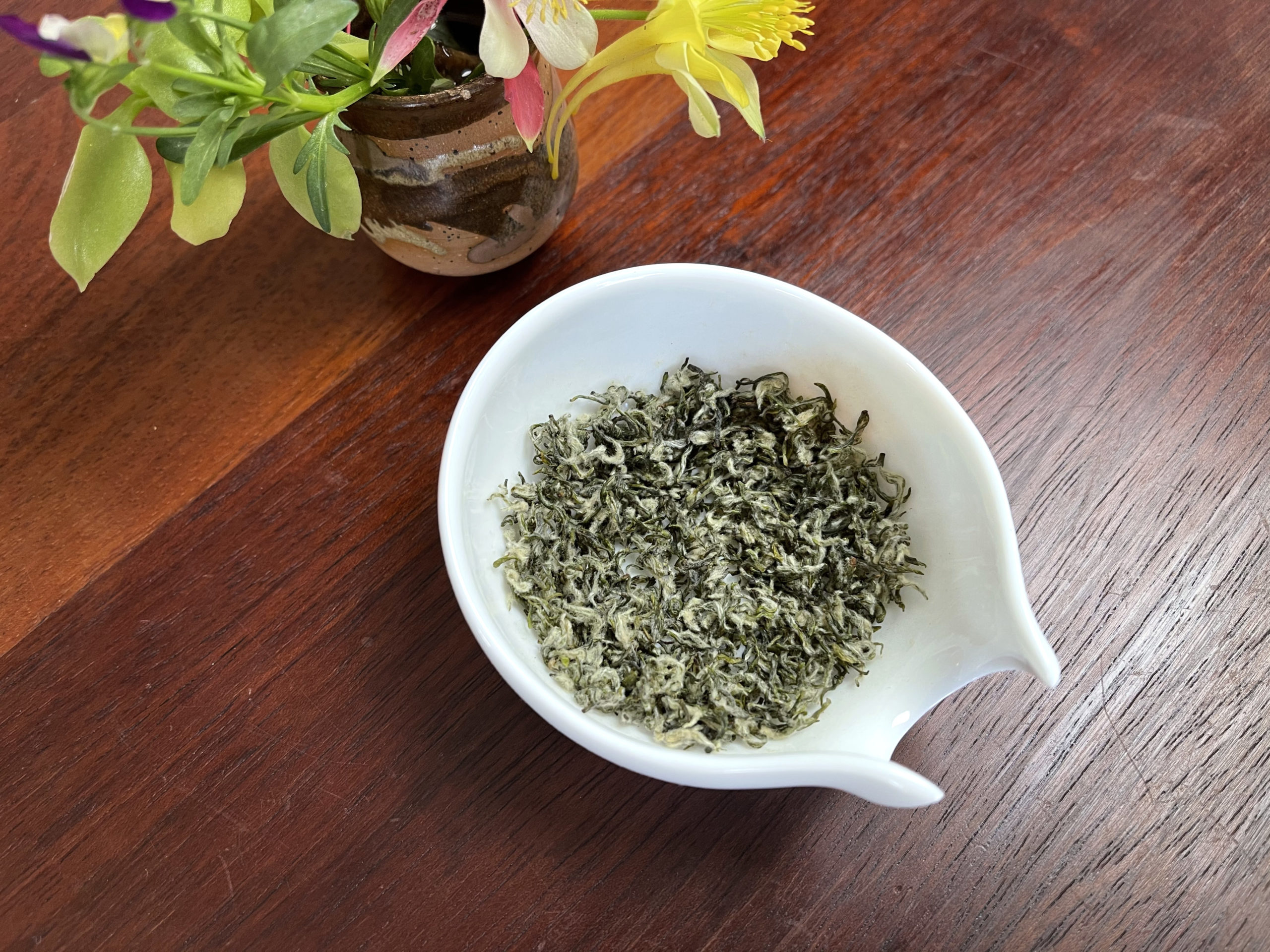 Very small and fluffy green tea leaves in a white dish on a woodent able next to a small flower bouquet.