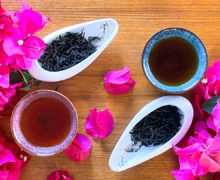 Close view from above of two wide porcelain cups of dark tea and two display dishes of dry black tea leaves, surrounded by fresh pink bougainvilleas.