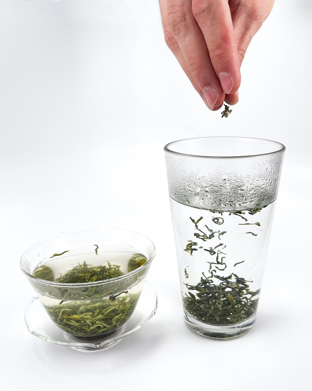 A hand sprinkling dry green tea leaves into a pint glass of hot water where they sink to the bottom and unfurl. Next to it, a glass gaiwan brewing already opened leaves.
