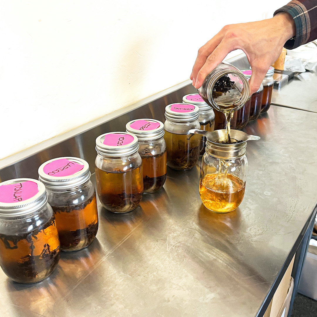 A row of canning jars full of cold brewing black tea. In front of them, someone pours out a jar into another jar through a strainer to catch the leaves.