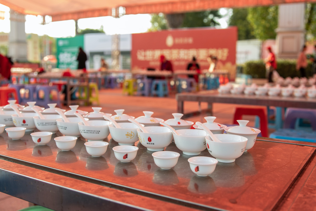 A row of gaiwan, cups, and bowls on a long steel table. Several other tables set up the same way and surrounded by stools are out of focus in the background. The whole area is under a large tent canopy.
