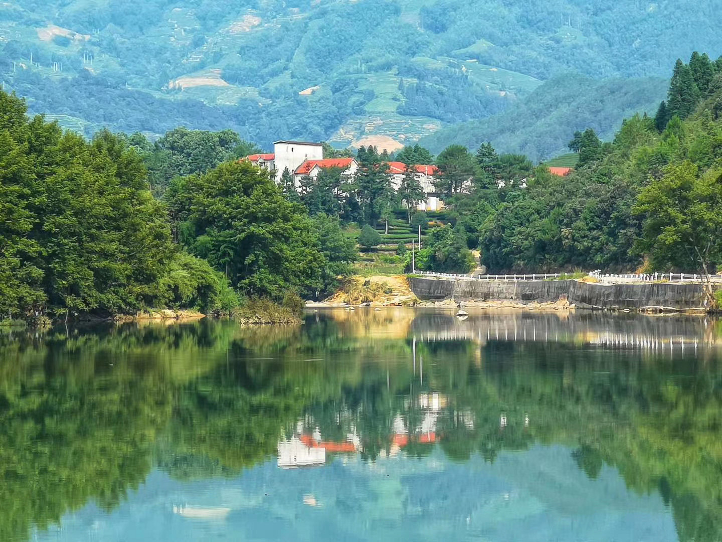 Looking across a river to the red roofs and white walls of a tea factory on a hillside half hidden by trees, surrounded by heavily forested hills and tea terraces.