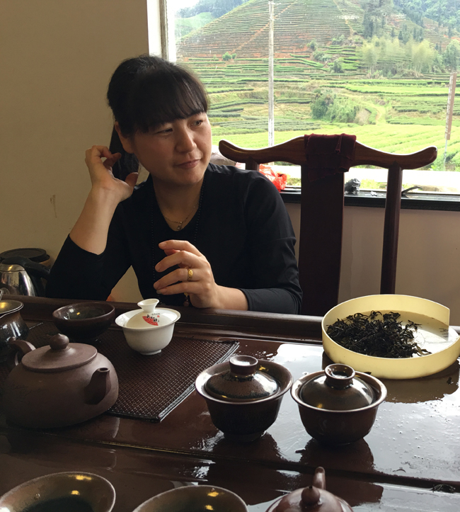 Huang Shiying leaning on a table while having tea in front of a window. There are rows of tea bushes on the hill visible outside.