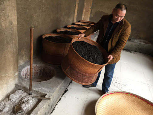 Tea maker Zhou Yousheng lifting a large basket of roasting rock wulong tea off a recessed charcoal stove to mix the leaves.