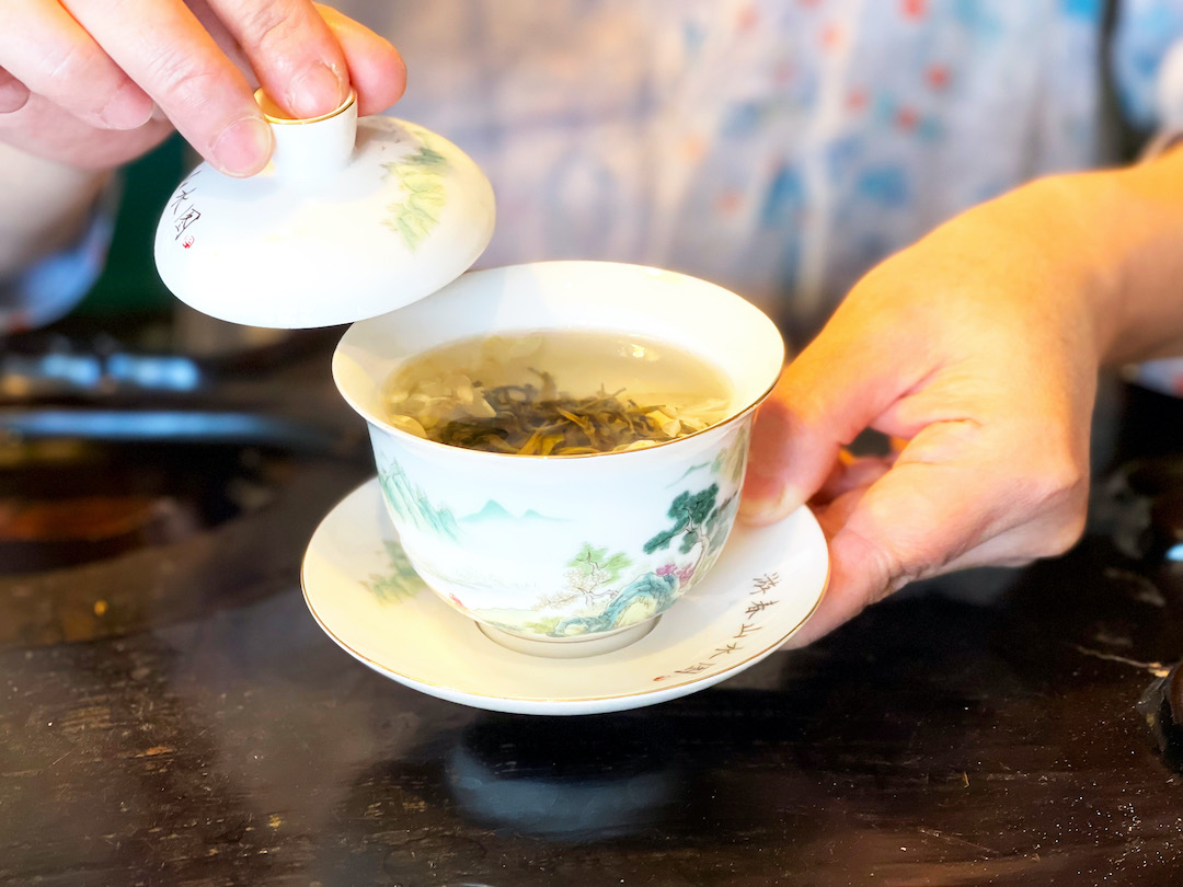 Close view of a painted white gaiwan held up in both hands to show the tea leaves and jasmine petals brewing inside, holding the lid open with one hand and bracing the saucer in the other.