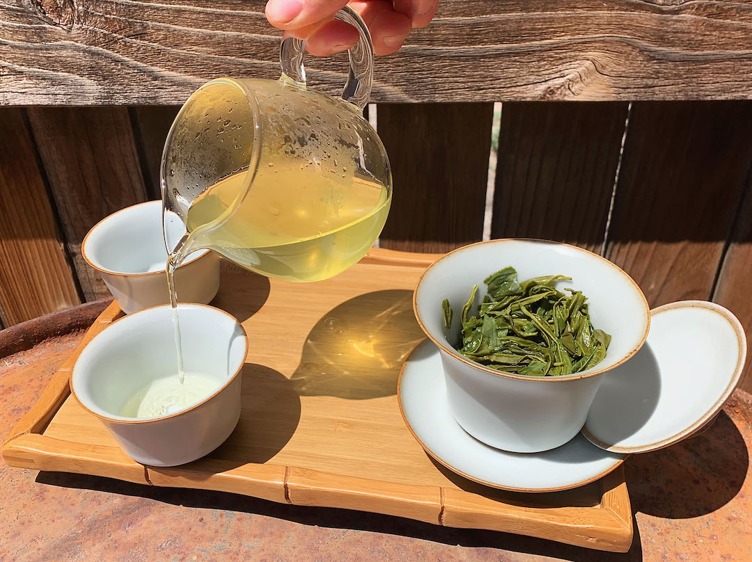 A leaf-filled gaiwan with the lid propped open between the cup and saucer, with two matching cups on a bamboo tray. A brightly sunlit glass pitcher pours green tea into a cup and throws sparkling shadows onto the tray.
