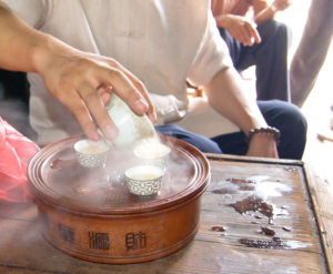 A hand pouring steaming hot tea out of a gaiwan into three small porcelain cups on a round clay tea tray.