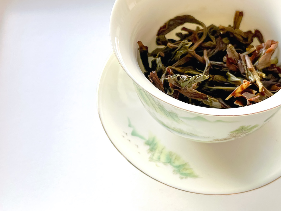 Colorful brown and green wulong tea leaves in an open white gaiwan with a subtle mountain pattern on the outside.