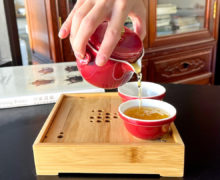 Close view of amber colored black tea pouring from a soft ruby red easy gaiwan into two matching cups. The cups sit on a bamboo tray and a book on Yixing teapots is visible in the background.