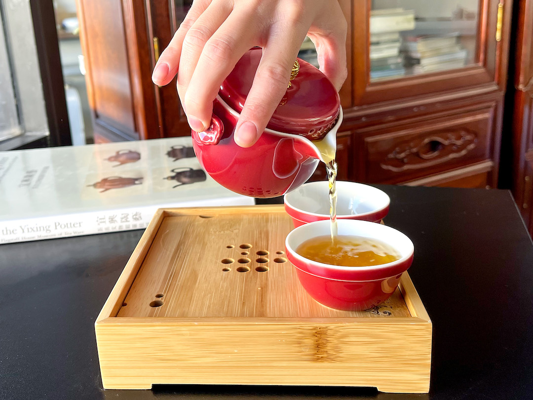 Close view of amber colored black tea pouring from a soft ruby red easy gaiwan into two matching cups. The cups sit on a bamboo tray and a book on Yixing teapots is visible in the background.