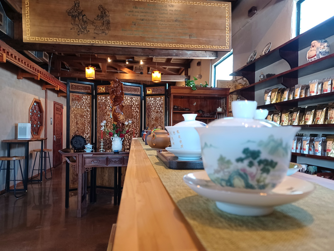 Looking down a shelf of teaware, focusing on a yixing teapot in the middle distance, a few unfocused gaiwan in the foreground, and the teahouse in the background, with a wooden statue of Guanyin visible in front of a carved wooden screen.