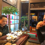Two people sitting at a table indoors drinking tea with a big spread of teaware between them.