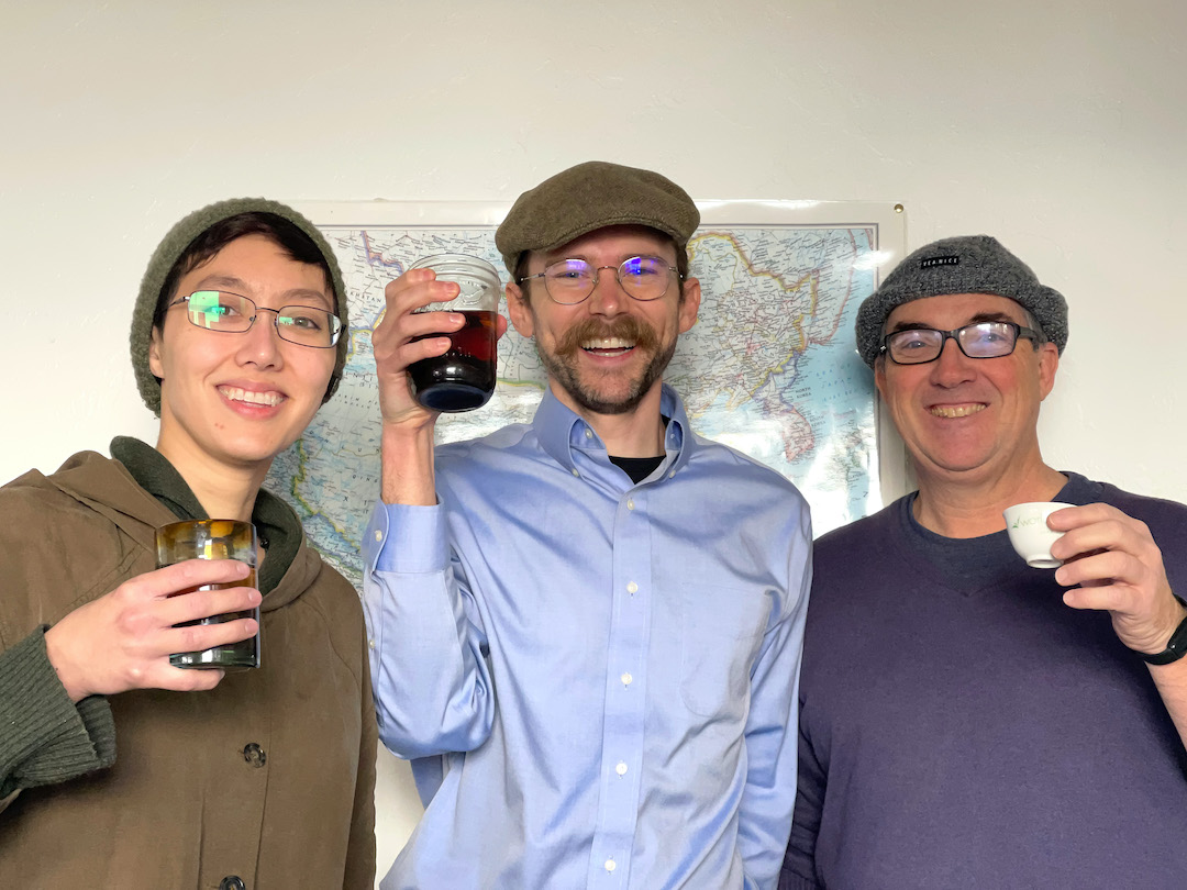 Three people with glasses and fabric hats lifting cups of tea and smiling at the camera.