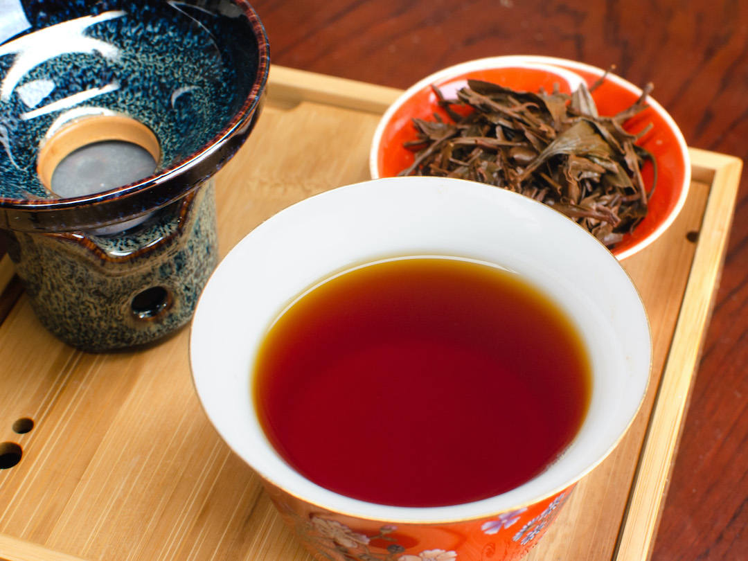 Close view of an open gaiwan full of the deep red infusion of a black tea, surrounded by a tea strainer and the inverted gaiwan lid full of strained leaves.