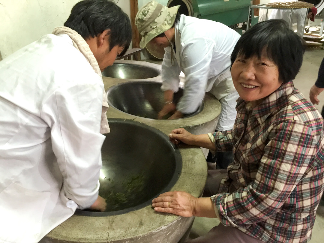A smiling woman seated next to a bank of adjacent frying woks supervising the two people leaning over them to fry Mogan Huangya yellow tea.