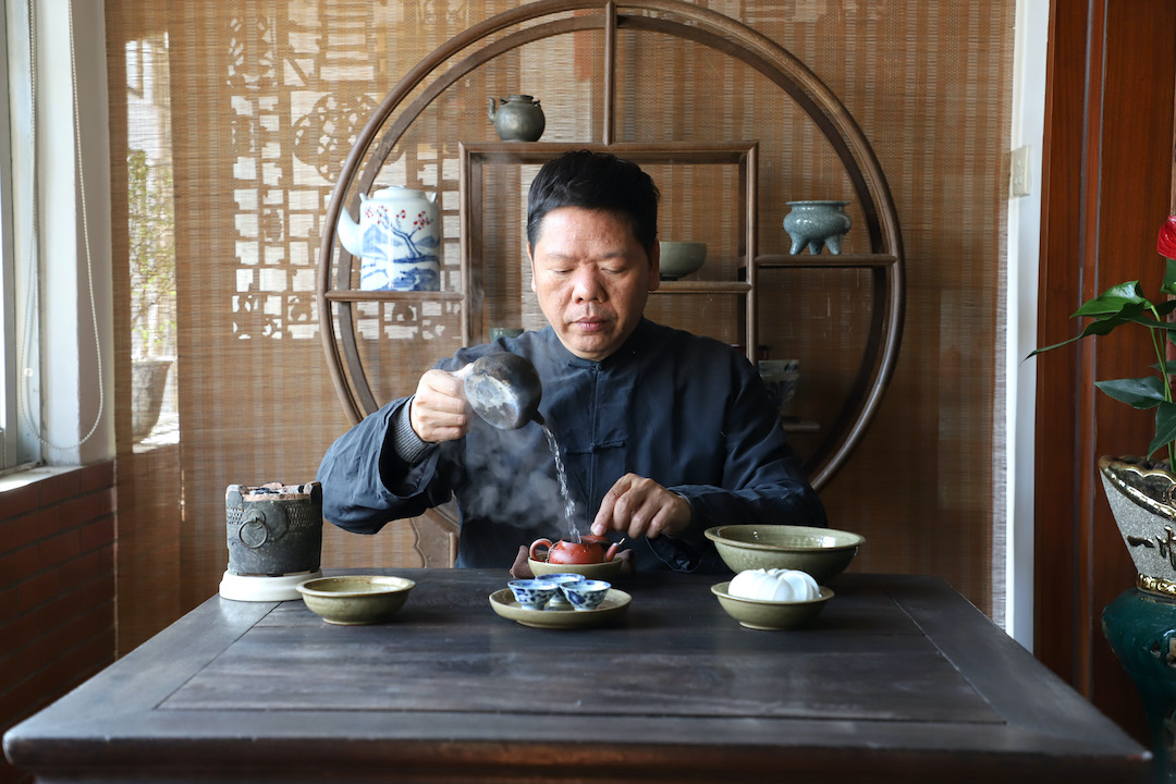 A man seated at a tea table brewing Dan Cong Wulong tea gongfu style, with an ornate shelf holding teaware behind him.