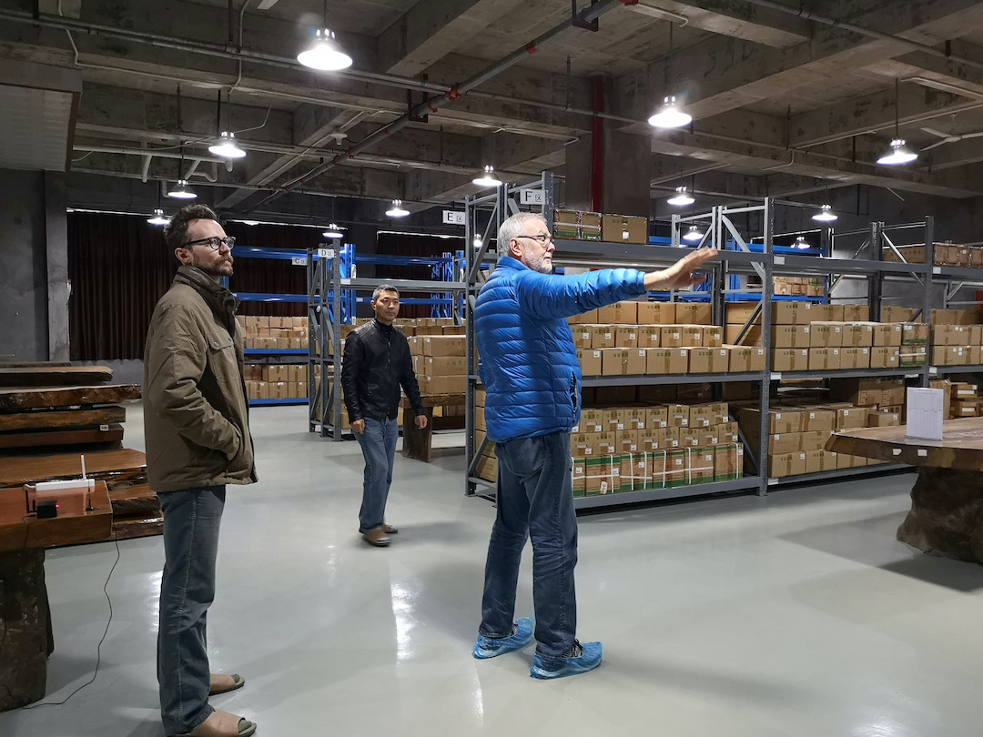 Three people standing at the end of several aisles of shelving lined with boxes in a well-lit warehouse for puer storage.