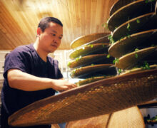 A man pulling a large round bamboo tray out from a rack of many others to check the tea leaves spread on it.