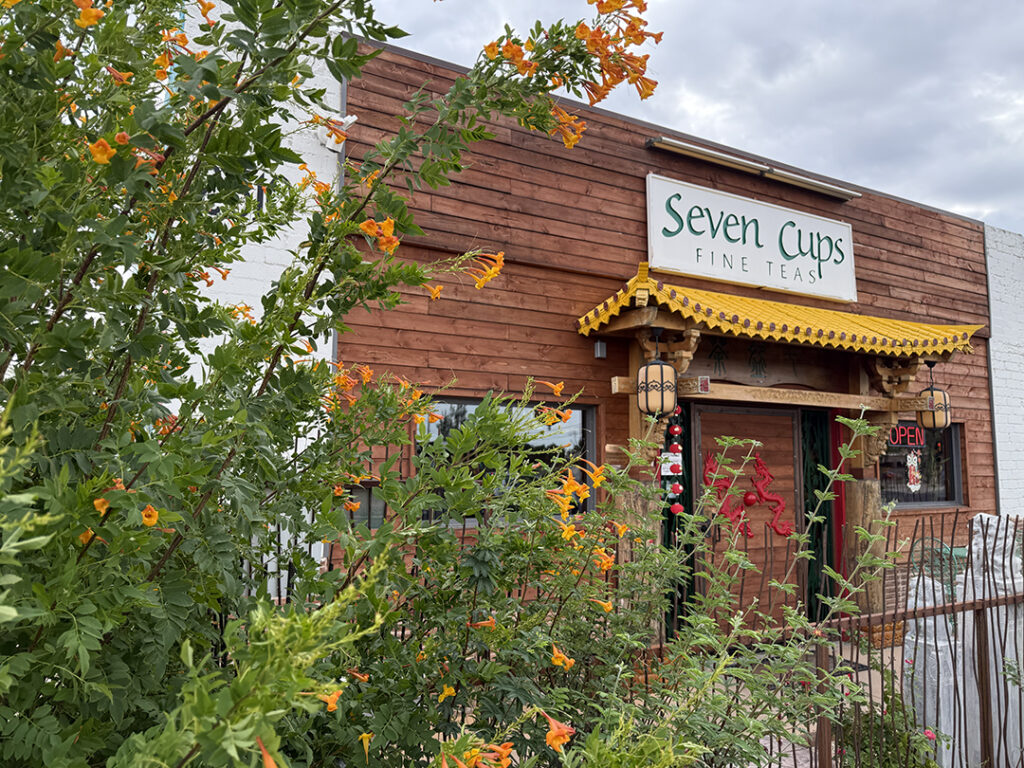Blooming shrubs enclose a white brick building with a carved wooden facade.