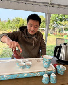 A man pouring tea from a dark red yixing pot into 4 bight azure blue gongfu tea cups.
