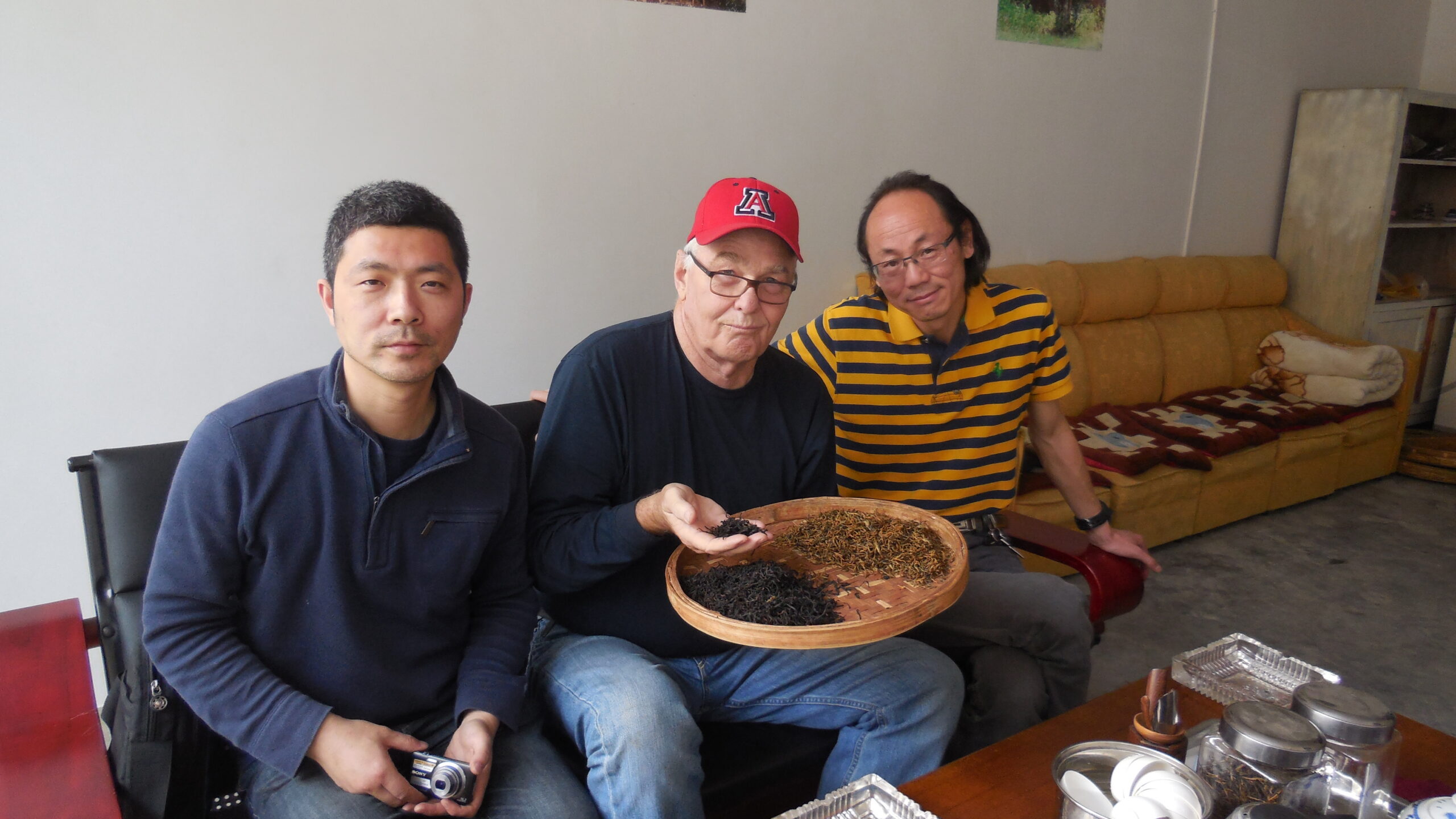 3 men sitting next to eachother looking at a basket of tea leaves. 