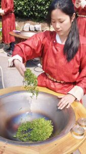 A woman in a bright red dress hand drying tea in a hot wok.