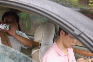 two men sitting in a car, one is looking at a flip phone and the other in the backseat is holding up a camera smiling taking a photo