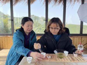 two women sitting next to eachother. The younger woman is pouring tea into the others cup with a chinese soup spoon and the tea leaves dance in a glass bowl.