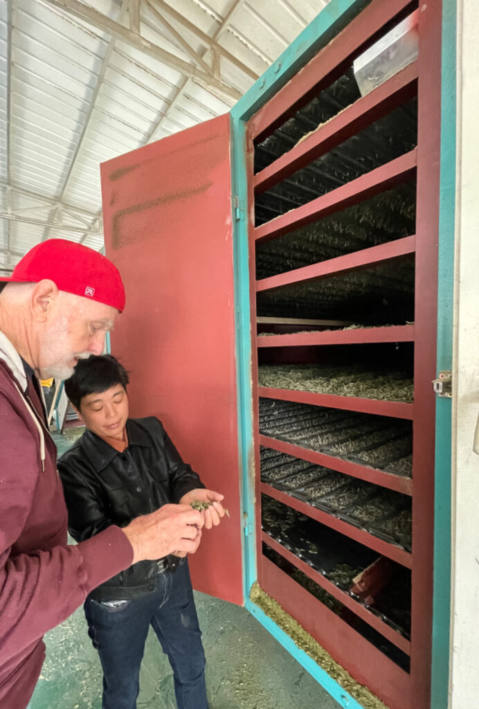 A woman in a leather jacket showing a man in a baseball cap the tea leaves being dried in a tall cabinet.