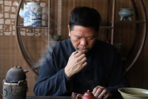 A man with his head bowed, smelling the tea from a small tea cup.