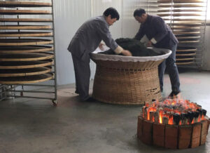 Two men mixing tea leaves in a large bamboo basket. on the floor is a fire that the basket is passed over to roast the tea.